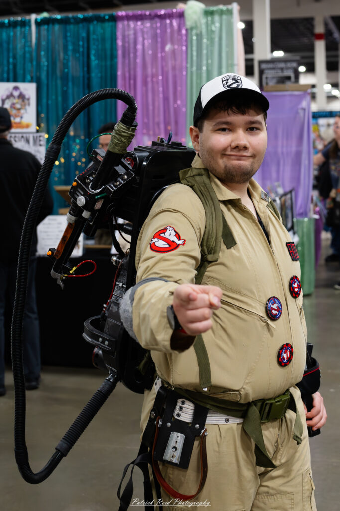"A cosplayer dressed as a Ghostbuster, complete with a tan jumpsuit, proton pack, and Ghostbusters logo patch, standing ready to battle supernatural foes."